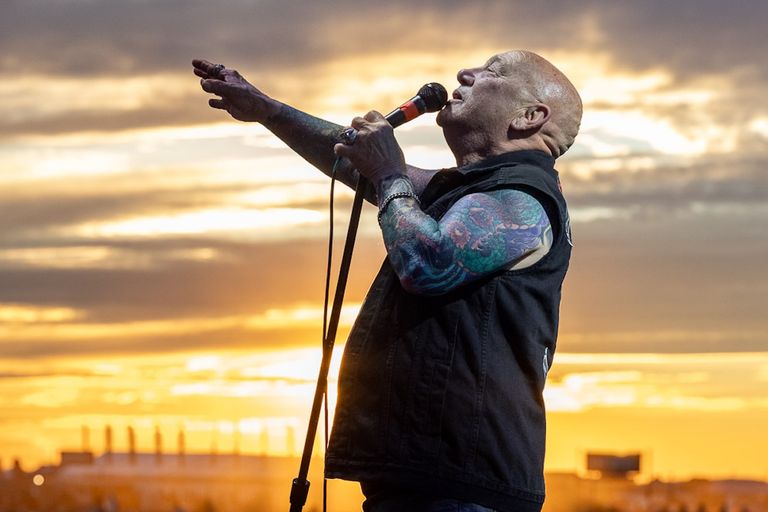 Rose Tattoo singer Angry Anderson singing into a microphone on an outdoor stage at sunset, reaching out to the audience.